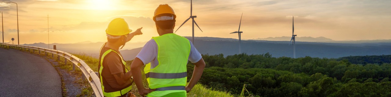 Two engineers looking at a wind power generator in front of the sun setting.